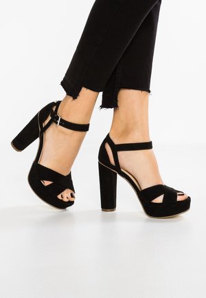 Žena na sobě má černé semišové vysoké sandály na podpatku s ankletovými pásky a otevřenými prsty, které jsou zkombinovány s černými zkrácenými kalhotami s třásněmi.