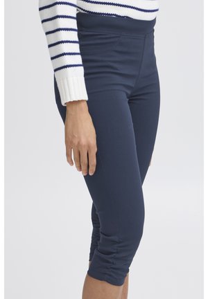 Žena v tmavomodrých krátkych legínach a bielej svetri s tmavomodrými pruhmi stojí pred jednoduchým svetlým pozadím.