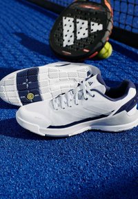 Bílé sportovní boty s námořnickými akcenty, síťovaným svrškem, zaoblenou špičkou a texturovanou podrážkou. V pozadí černý pádlo a tenisový míček.