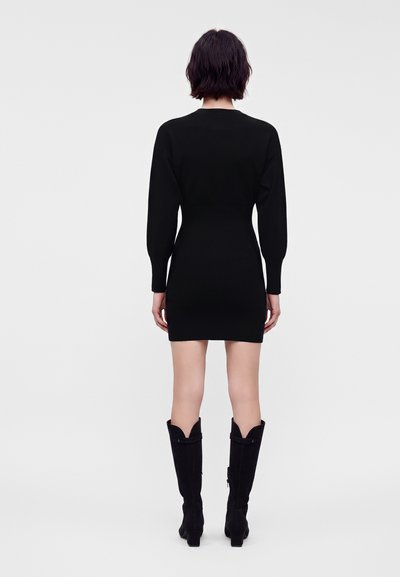 Femme aux cheveux courts et foncés, portant une mini-robe noire à manches longues et des bottes noires montantes, debout de dos sur un fond uni.