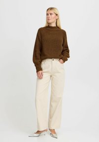 Pulover tricotat maro cu guler înalt, textură ribbed; pantaloni crem cu picioare largi; pantofi argintii cu vârful ascuțit și finisaj lucios.