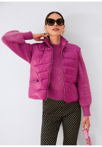 Aufgeblasene Weste in leuchtendem Lila mit Stehkragen, kombiniert mit einem passenden lila Pullover und schwarzen gemusterten Hosen mit weißen Quadraten.