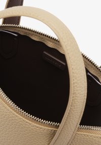 Borsa a mano beige con texture e chiusura a zip, con interni marroni e una piccola etichetta in pelle del marchio all'interno. Due manici corti visibili.