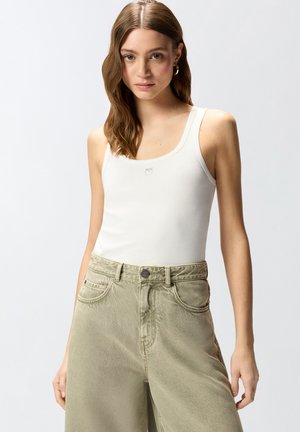 Mladá žena s dlouhými vlnitými vlasy, která nosí zlaté kruhové náušnice, bílý top bez rukávů a světle olivové kalhoty s vysokým pasem a širokými nohavicemi.