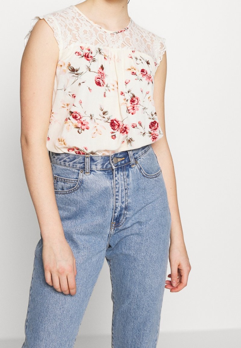 Blusa floral con un yugo de encaje, tela suave en color crema, rosas rosas y rojas, corte holgado, combinada con jeans de mezclilla azul de cintura alta.