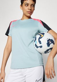Camiseta deportiva Nike de color azul claro con mangas rosas y negras, combinada con pantalones cortos blancos. El jugador sostiene un balón de fútbol de diseño.