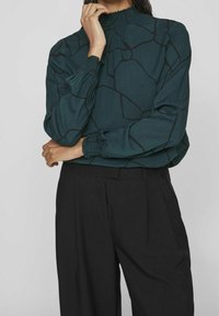 Blusa verde con colletto alto e maniche lunghe, caratterizzata da un motivo geometrico nero, polsini arricciati e un tessuto fluido. Abbinata a pantaloni neri.