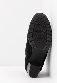 Chaussure noire à talon avec semelle en caoutchouc texturée et talon bloc, positionnée droite contre un coin blanc.