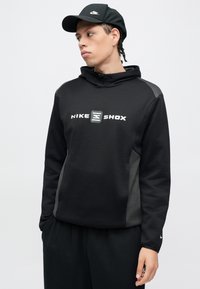 Μαύρο φούτερ Nike με λογότυπο μπροστά, γκρι πλευρικές πινελιές, κουκούλα με κορδόνια και μακριά μανίκια. Φορώντας με ταιριαστό μαύρο παντελόνι και καπέλο.