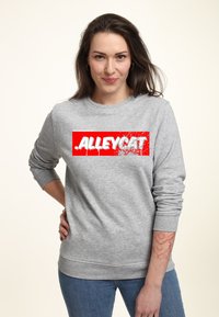 Šedá mikina s výrazným červeným potiskem "ALLEYCAT". Má kulatý výstřih a žebrované manžety, kombinováno s modrými džíny.