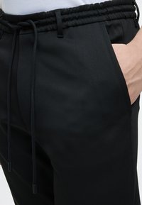 Personne portant un pantalon noir à cordon avec une main dans la poche droite, montrée de la taille au milieu de la cuisse sur un fond uni.