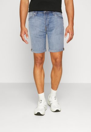 Homme portant un short en jean bleu clair retroussé, des chaussettes blanches et des baskets blanches, debout devant un fond uni clair.