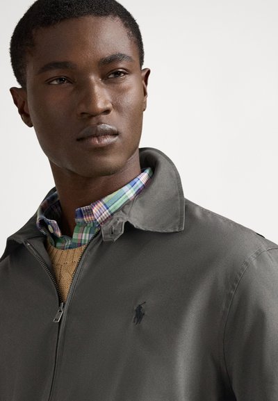 Chaqueta gris oscuro con cremallera y cuello, que se lleva sobre una camisa de cuadros. La chaqueta presenta un pequeño logotipo bordado en el pecho.