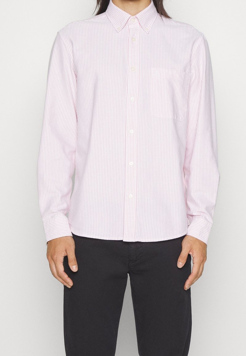 Chemise à rayures roses claires avec boutons, col, manches longues et poche poitrine. Fabriquée à partir d'un tissu lisse. Portée avec un pantalon foncé.