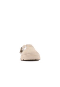 Beige mockaskinnsko med en formad gummisula, stängd tå och justerbar remdetalj på sidan, med slät yta och rena linjer.