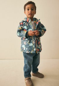 Giacca impermeabile con base blu-verde arricchita da motivi colorati di animali e natura, chiusura con zip e cappuccio foderato. Indossata con jeans di denim.