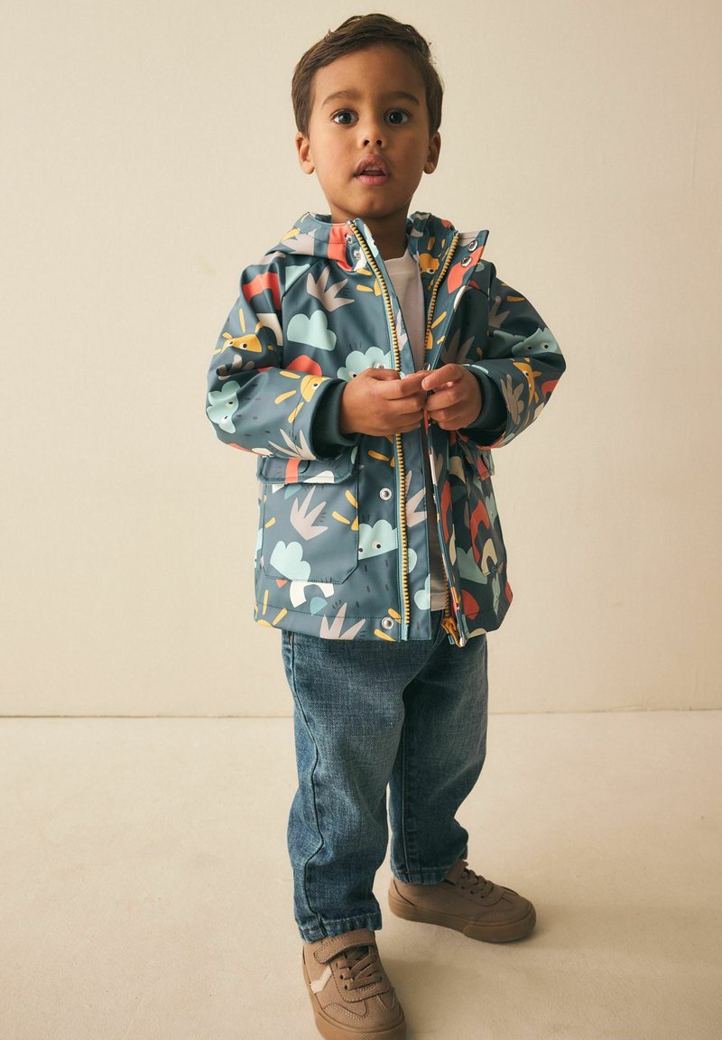 Giacca impermeabile con base blu-verde arricchita da motivi colorati di animali e natura, chiusura con zip e cappuccio foderato. Indossata con jeans di denim.