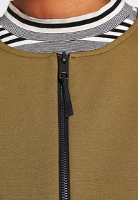 Olivgrön zippered plagg med en svart dragkedja, lagd över en svart-och-vit randig polotröja. Slät textur, avslappnad design.