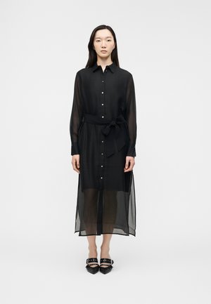 Femme portant une robe longue noire transparente boutonnée avec une ceinture nouée à la taille et des chaussures noires à bouts pointus avec des lanières et des détails métalliques.