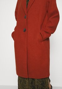 Long manteau couleur rouille avec des boutons noirs porté sur une jupe à motifs verts et noirs, mains dans les poches du manteau.