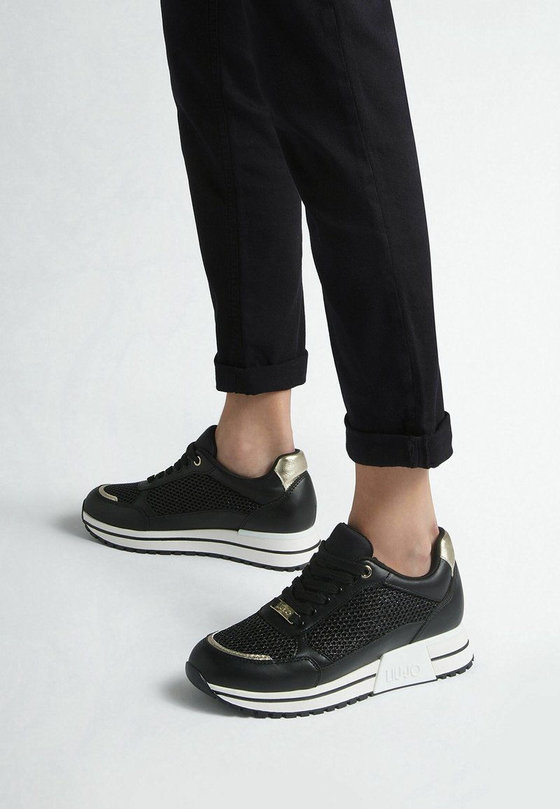 Schwarze Sportschuhe mit einem Obermaterial aus Mesh, goldenen Akzenten, runden Schnürsenkeln und einer gestapelten weißen Sohle, kombiniert mit hochgekrempelten schwarzen Hosen.