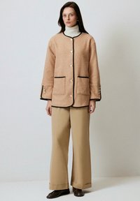 Giacca beige testurizzata con rifiniture nere, tasche frontali e grandi bottoni, abbinata a pantaloni beige larghi e un dolcevita bianco.