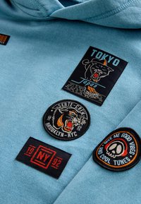 Světle modrá mikina s kapucí, která obsahuje několik vyšívaných nášivek: černý panter, "Tokio," "Skate Crew Brooklyn" a kulatou nášivku "Dobré Vibe".