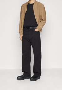 Veste bomber beige, chemise noire, pantalon ample noir, ceinture noire et chaussures noires imposantes. Matériaux texturés avec des poignets élastiques.