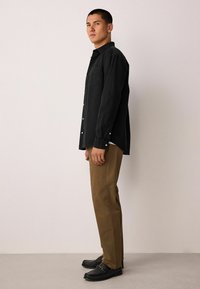 Mustast nööpidega särk rinnataskuga, kombineeritud beežide pükste ja mustade mokassiinidega, seismas ühevärvilise tausta ees.