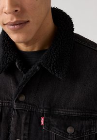 Zwarte denimjas met een gestructureerde zwarte sherpa kraag, sluiting met knopen aan de voorkant en een rode Levi's-label op de borstzak.