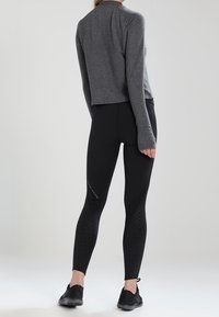 Grijze cropped longsleeve top gecombineerd met zwarte leggings met textuurpatronen en contrasterende accenten, gedragen met zwarte sportschoenen.