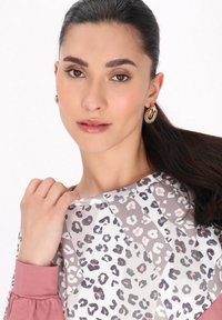 Sötét hajú nő, akinek a haja hátra van fogva, arany karika fülbevalót visel és rózsaszín leopárdmintás felsőt hord, az egyik kezével gyengéden a gallérját fogja.