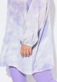 Tunika leggera in tinta unita lavanda e bianca, con maniche lunghe e polsini elasticizzati, e orlo arrotondato. Abbinata a leggings coordinati.