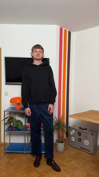 Fiatal férfi fekete kapucnis pulóverben és sötét nadrágban áll egy szobában, melyben televízió, narancssárga lámpa, cserepes növények, színes falcsíkok és egy fém bőrönd található.