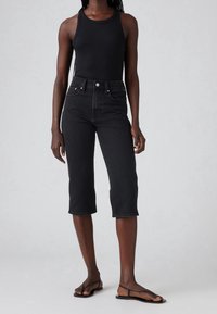 Jeans negros recortados con una cintura alta, que presentan un diseño clásico de cinco bolsillos y costuras en contraste. Combinados con una camiseta sin mangas de canalé negra y sandalias planas.