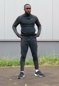 Schwarzer Sportanzug mit langen Ärmeln, verziert mit gemusterten Akzenten an Armen und Beinen, kombiniert mit schwarzen Sportschuhen und weißen Socken.