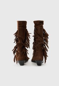 Bottes en suede marron jusqu'au genou avec des franges sur les côtés et un petit talon block. Texture lisse et ajustement confortable autour du mollet.
