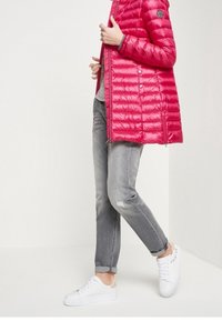 Heldere roze, lichtgewicht donsjas met ritssluiting, voorzien van zijzakken en geribde manchetten. Draagt grijze spijkerbroek en witte sneakers.