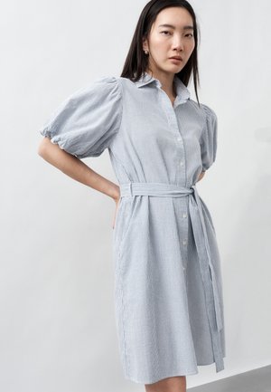 Mujer con un vestido a rayas azul celeste y blanco con cinturón y mangas cortas abullonadas, de pie con una mano en la cadera sobre un fondo liso.