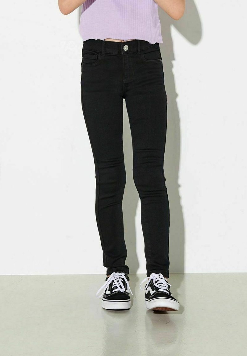 Personne portant un jean skinny noir, des baskets noires et blanches, et un top court en maille côtelée lavande, debout devant un fond blanc.