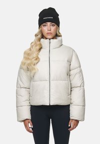 Hellbeige Pufferjacke mit hohem Kragen, Reißverschluss vorne und gerippten Bündchen. Mit auffälligem weißen Branding auf der linken Brust.