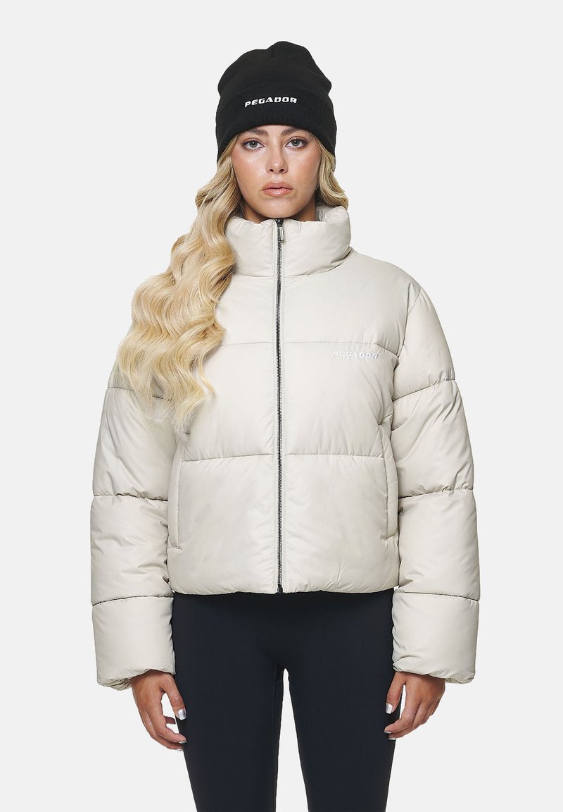 Hellbeige Pufferjacke mit hohem Kragen, Reißverschluss vorne und gerippten Bündchen. Mit auffälligem weißen Branding auf der linken Brust.