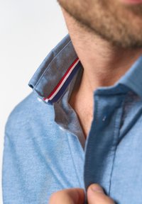 Light blue polo shirt with a folded collar featuring red, white, and blue stripes inside. Soft texture and casual design, partially unbuttoned.