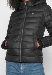 Czarna ocieplana kurtka z zamkiem z przodu, elastycznymi mankietami i kapturem. Ma poziome przeszycia i detal z logo. Gładka tekstura.