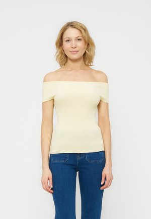 Mujer rubia con un top de canalé color crema con los hombros descubiertos y pantalones vaqueros azules, posando frente a un fondo blanco liso.