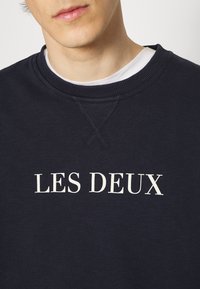 Sudadera azul marino con cuello redondo acanalado y texto blanco "LES DEUX" impreso en el frente. Tela suave con textura lisa.