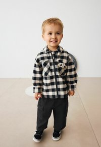 Un jeune enfant souriant, portant une veste noire et blanche à carreaux avec un patch de panda, un pantalon noir et des baskets noires, se tenant à l'intérieur sur un sol beige.