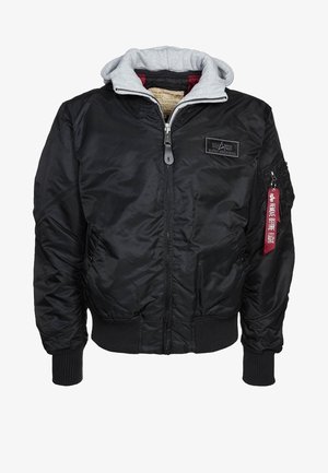 Veste bomber noire avec capuche grise, fermeture éclair à l'avant, poches zippées, poignets et ourlet côtelés, et étiquette rouge "Remove Before Flight" sur la poche de la manche gauche.