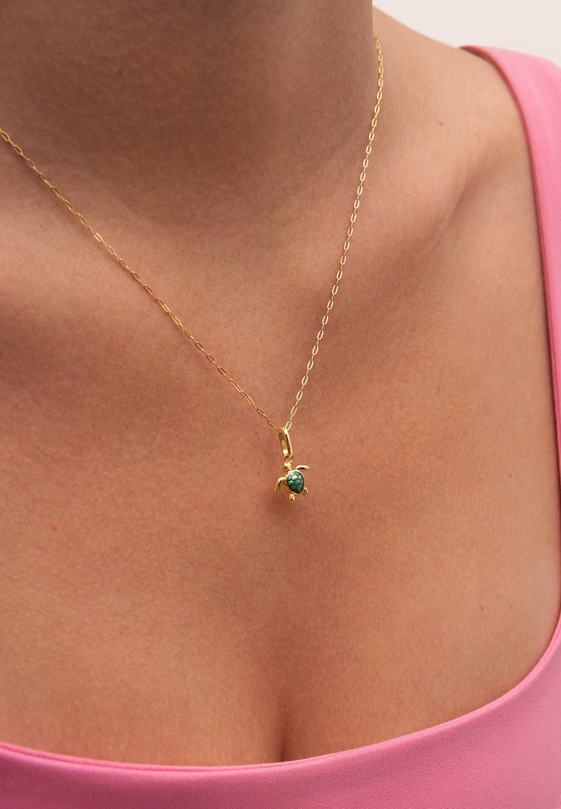 Collar de cadena de oro con un colgante de tortuga y un acento verde. Llevado en un cuello desnudo, sobre una blusa rosa. Textura suave y brillante.