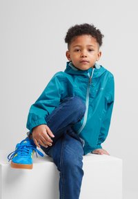 Teal lightweight jacket with a hood, zippered front, and elastic cuffs. Blue suede sneakers with yellow accents. Sitting on a white pedestal.